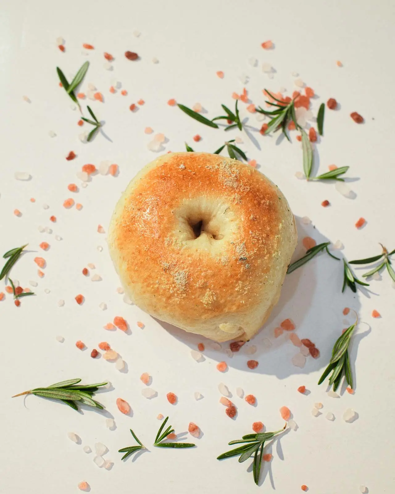 A rosemary salt bagel surrounded by fresh rosemary and coarse salt.