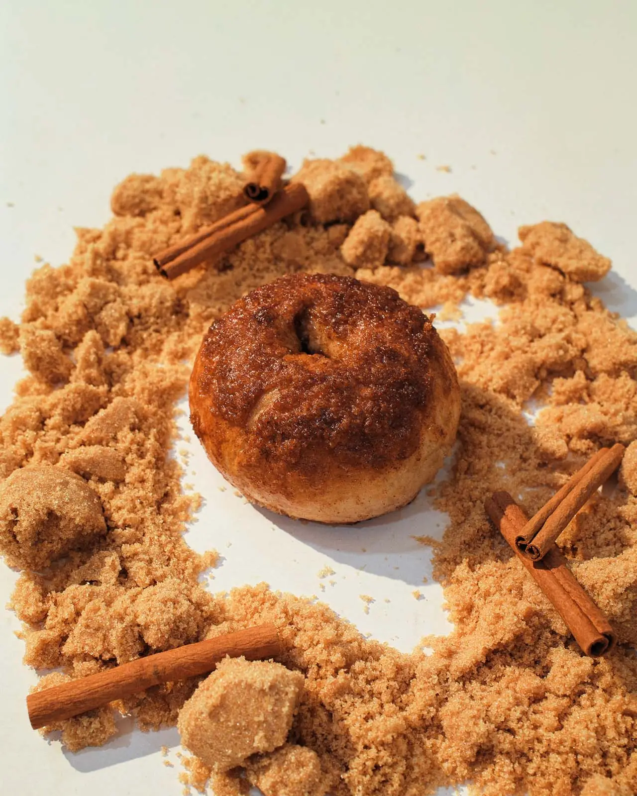 A cinnamon raisin bagel with a dark sugar crust, resting on loose brown sugar and cinnamon sticks.