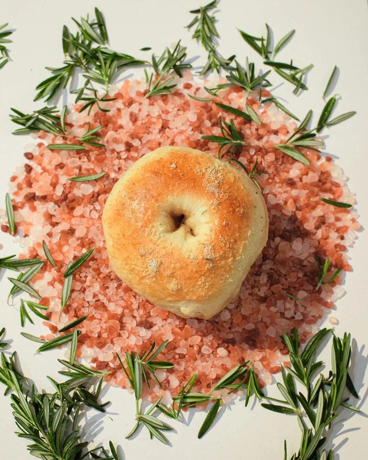 A rosemary salt bagel on a bed of pink rock salt with sprigs of fresh rosemary.