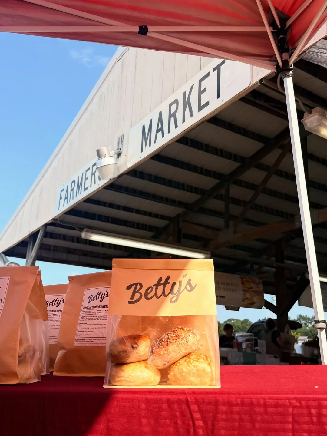 The Betty's Bagels stall at the Florence Farmers Market — branded kraft bags of bagels on a red table under the market sign.